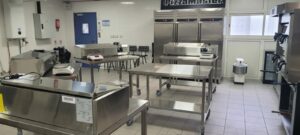 Photographie d'une cuisine professionnelle illustrant le principe de la marche en avant avec des zones sales et propres délimitées au sol par des flèches. Un chef travaille à une station de préparation propre, et une tablette au premier plan affiche le logo OAFormation avec une checklist d'audit de cuisine, soulignant l'importance de la réglementation et de l'ergonomie