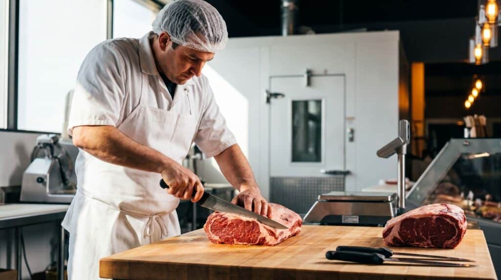 Boucher artisan en tenue de travail découpant une pièce de viande sur un billot propre dans un laboratoire de boucherie